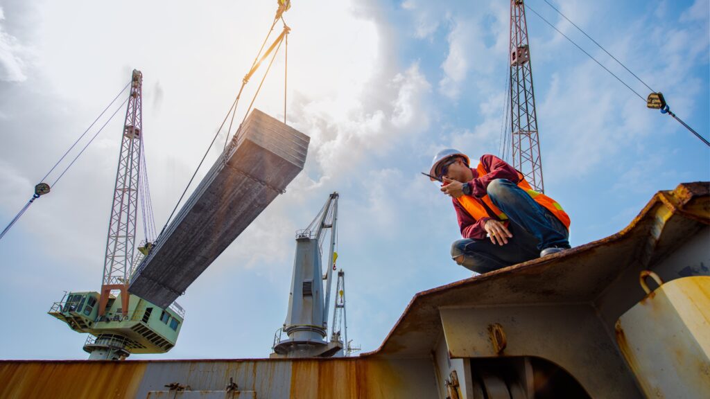 A construction worker with a walkie-talkie coordinates the positioning of a large piece of steel being delivered to a port